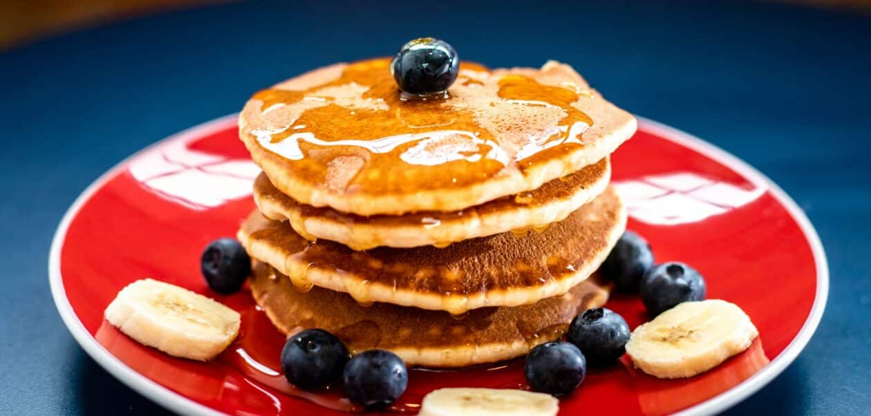 Stack of fluffy eggless pancakes topped with maple syrup and blueberries, served with banana slices on a red plate.