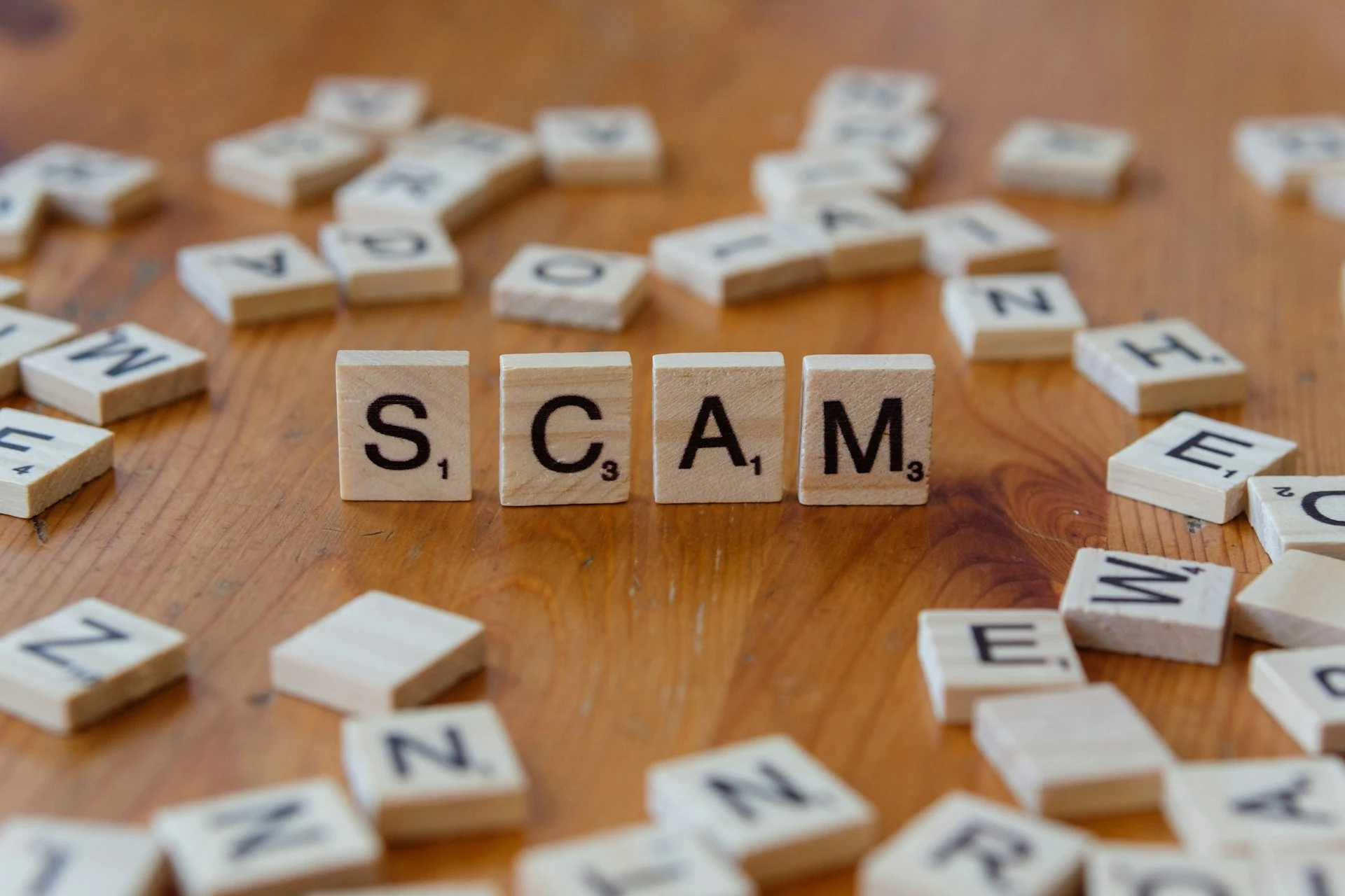 AI Refund Scams: Scrabble tiles on a wooden table spelling out the word "SCAM." Illustrates the intentional and costly nature of AI Refund Scams in quick commerce.